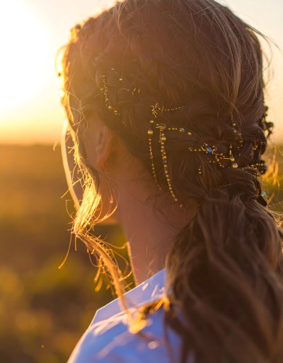 Woman's braided hair with gold beads and blue gems, backlit by sunset.