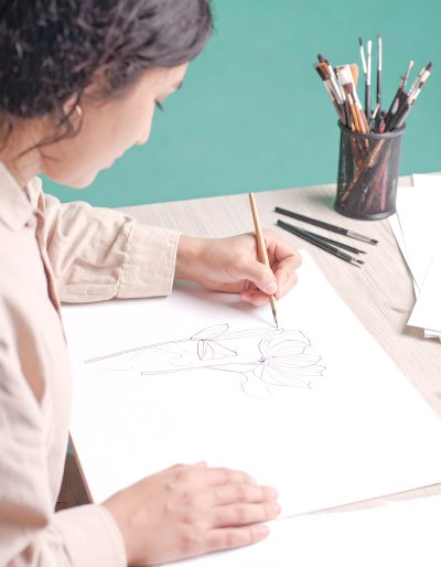 Artist sketching a flower with a brush, surrounded by art supplies.